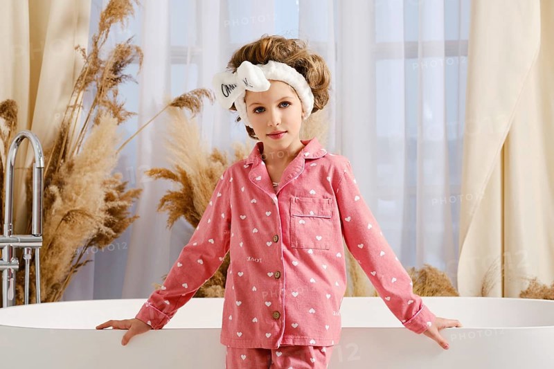 Cute child in a pink pajama and hair band stands next to a modern white bathtub, surrounded by elegant curtains and dried pampas grass