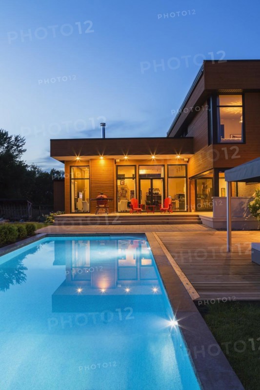 Rear of modern cube style home with stained horizontal wood cladding, in-ground swimming pool and wooden deck with gazebo canopy at dusk, Quebec, Canada, North America