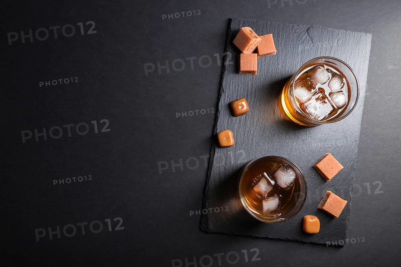 Glass of amber whiskey with ice and caramel candies on a black stone slate board on black background. Top view, copy space, flat lay, low key concept