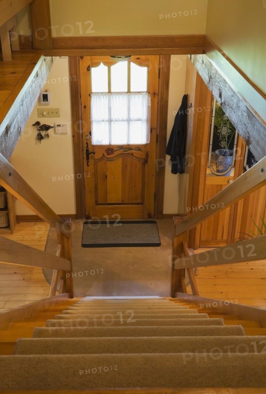 Steep staircase with tan carpet covered steps leading to downstairs floor and main entrance door inside Canadiana style log cabin home, Quebec, Canada, North America