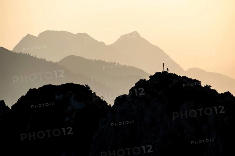 Lone mountaineer on the summit of the Hörndlwand in the Chiemgau Alps with the silhouette of Zwiesel and Hochstaufen against the light in the early morning, Bavaria, Germany, Europe
