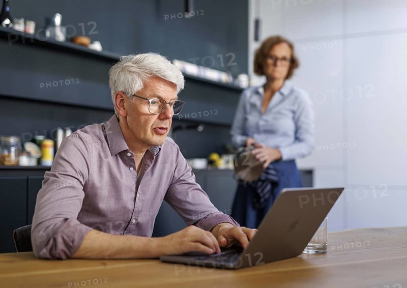 Symbolic photo on the subject of division of labour for couples in the household. A man sits in a kitchen at his laptop while a woman washes dishes in the background. Berlin, 13.08.2024