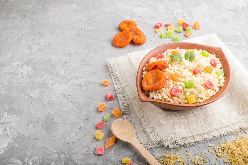 Bulgur porridge with dried apricots and candied fruits in clay bowl on a gray concrete background and linen textile. Side view, copy space. Turkish traditional cuisine