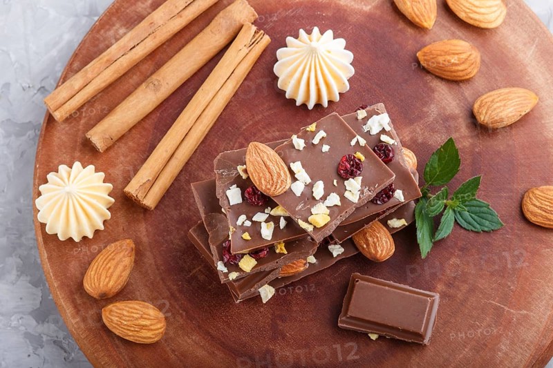 A pieces of milk chocolate with almonds and dried fruits on a brown wooden board on a gray concrete background. top view, close up, selective focus, flat lay