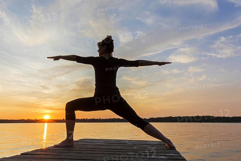Woman practicing yoga posture Virabhadrasana II, Warrior Pose, lunging standing asana on jetty at lake at sunset