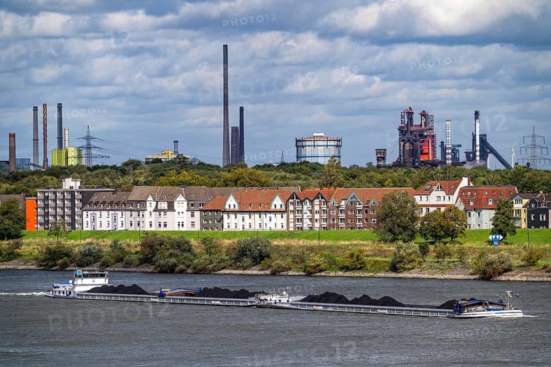 Cargo ship loaded with coal, on the Rhine near Duisburg-Laar, houses on Deichstraße, industrial backdrop of the ThyssenKrupp Steel steelworks in Bruckhausen, North Rhine-Westphalia, Germany, Europe