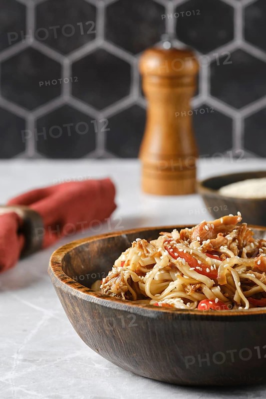 A flavorful dish of stir-fried noodles with colorful vegetables is presented in a rustic wooden bowl. The backdrop features a stylish hexagonal tile pattern, creating a cozy atmosphere