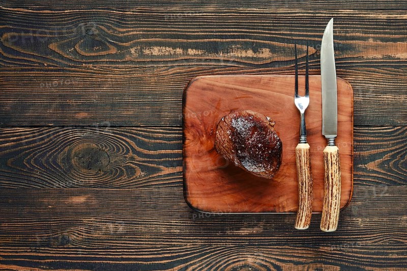 A beautifully grilled steak rests on a rustic wooden cutting board, accompanied by a knife and fork. The warm tones of the wood contrast with the deliciously caramelized meat, inviting enjoyment