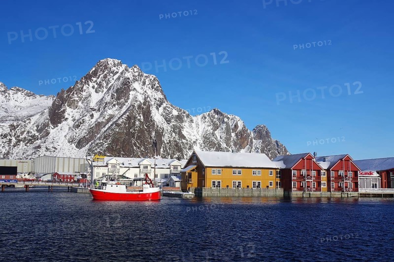 Colourful houses and a red fishing boat in the harbour in front of snow-covered mountains under a clear blue sky, Winter, Hotels, Svolvaer, Svinoya, Lofoten, Nordland, Norway, Europe