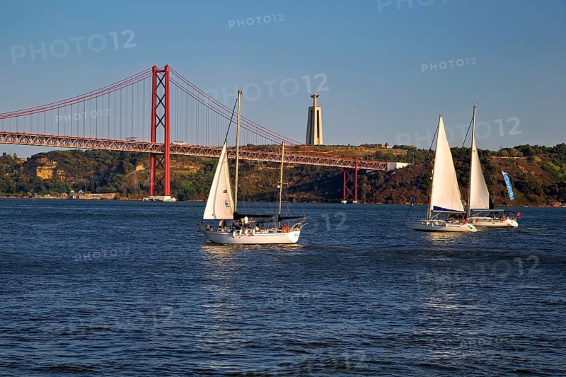 Two sailboats on calm waters with a red suspension bridge in the background under a blue sky, Lissabon, 25 de Abril Bridge