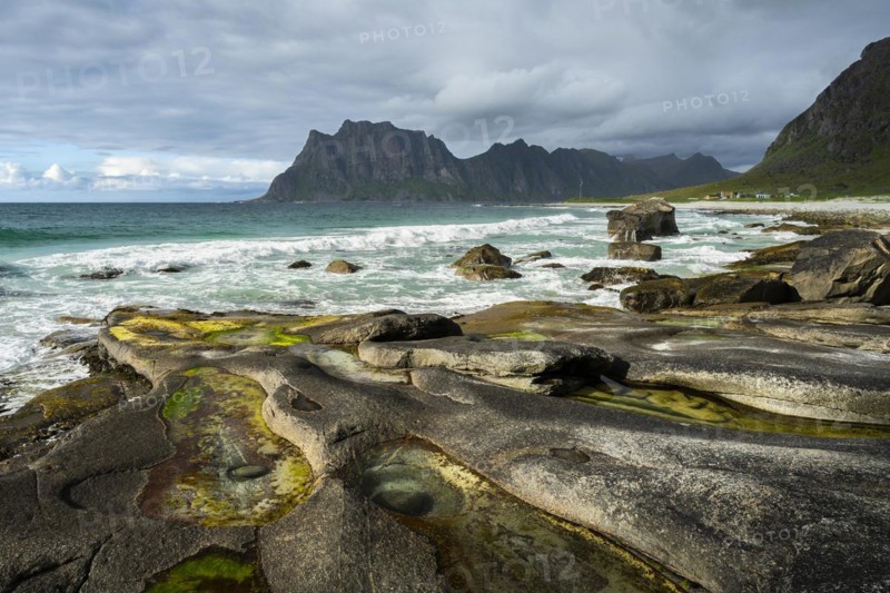 Seascape on the beach at Uttakleiv (Utakleiv), in the foreground rocks and rocky outcrops filled with water, including the so-called Eye of Uttakleiv, a round stone in the water. Mount Hogskolmen in the background. Sun and clouds. Early summer. Uttakleiv, Vestvagoya, Lofoten, Norway, Europe