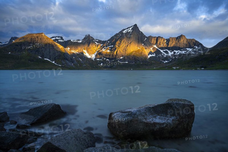Landscape with sea and mountains on the Lofoten Islands, view over the fjord Flakstadpollen to the mountain Stortinden and others. A rock in the foreground. At night at the time of the midnight sun, some clouds in the sky, the sun shining on the mountains. Long exposure. Early summer. Flakstadoya, Lofoten, Norway, Europe