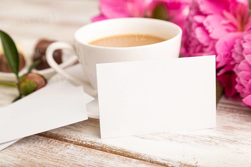 White business card with pink peony flowers and cup of coffee on white wooden background. side view, copy space, still life. Breakfast, morning, spring concept