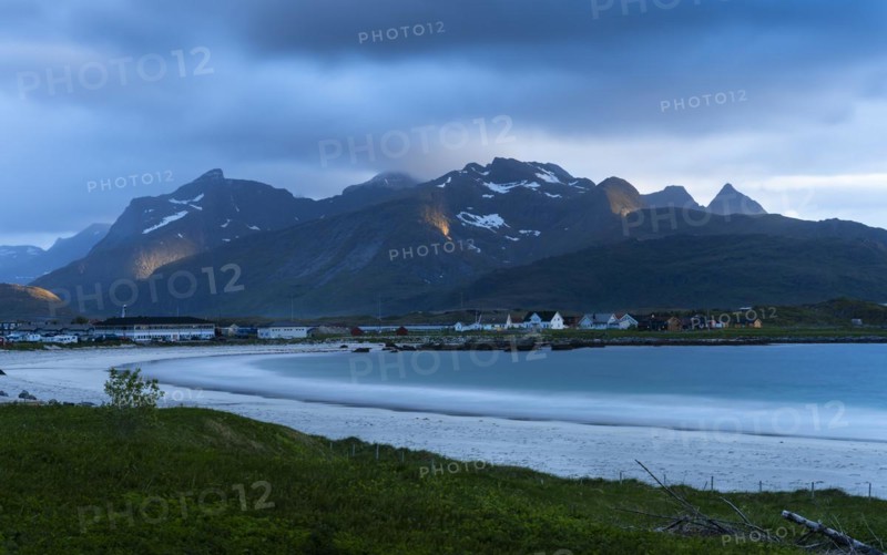 The village of Ramberg with sea and sandy beach. Mountains behind it. Individual sun spots in the mountains. At night at the time of the midnight sun. Clouds in the sky. Early summer. Long exposure. Ramberg, Flakstadoya, Lofoten, Norway, Europe