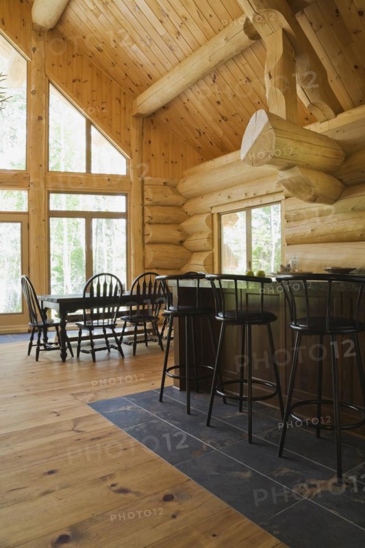 Island with dark grey granite countertop and black stained wooden bar stools plus antique table with rounded high back chairs in open concept kitchen dining room inside handcrafted Eastern white pine Scandinavian log cabin home, Quebec, Canada, North America