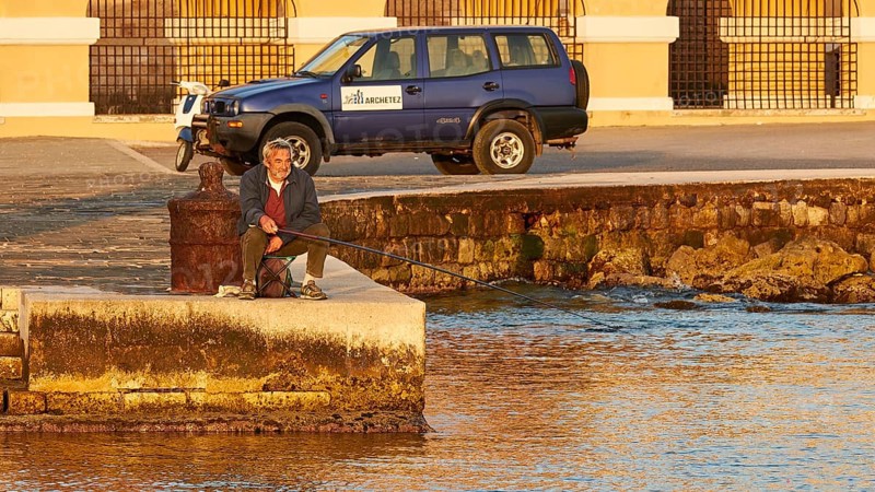 A man relaxes while fishing by the sea next to a parked off-road vehicle, Mandraki Oat, harbour area, Rhodes Town, Rhodes, Dodecanese, Greek Islands, Greece, Europe