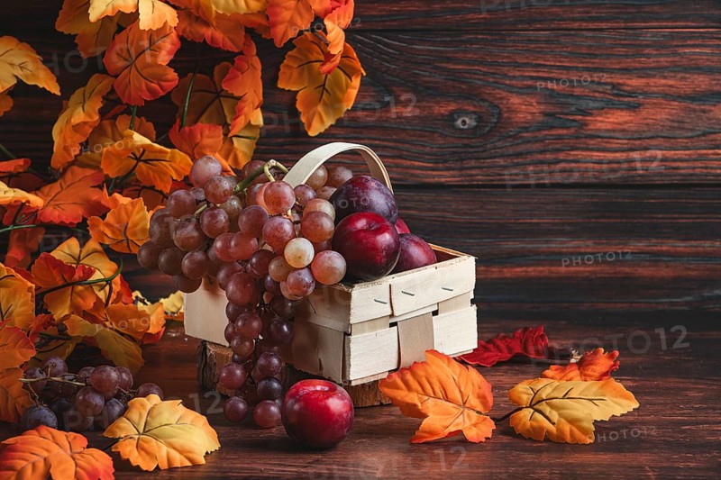 Grapes and plums in a wooden basket surrounded by autumn leaves on a table