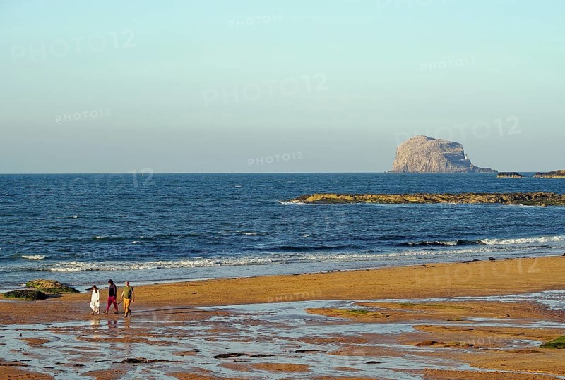 Walkers on the beach with rocks and waves in the background under a clear sky, Bass Rock, North Berwick, Scotland, United Kingdom, Europe