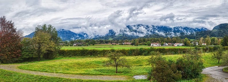 Wide landscape with green meadows, trees and a village in front of a mountain range under a cloudy sky, Bad Feilnbach