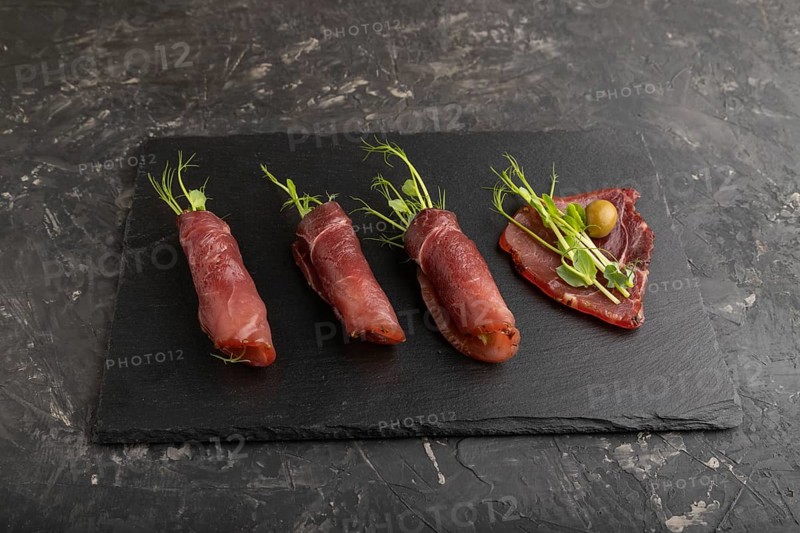 Slices of smoked salted meat with green pea microgreen on black concrete background. Side view, close up