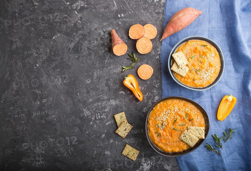Sweet potato or batata cream soup with sesame seeds and snacks in blue ceramic bowls on a black concrete background with blue linen textile. top view, flat lay, copy space