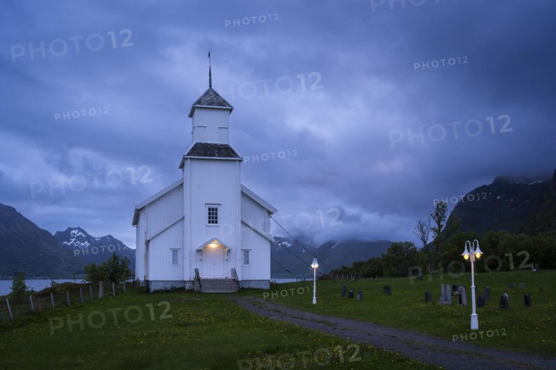 The white church of Gimsoy (Gimsoysand church) by the sea. A path leads to the church. The church and lanterns are illuminated. At night at the time of the midnight sun. Cloudy sky. Early summer. Gimsoya, Lofoten, Norway, Europe