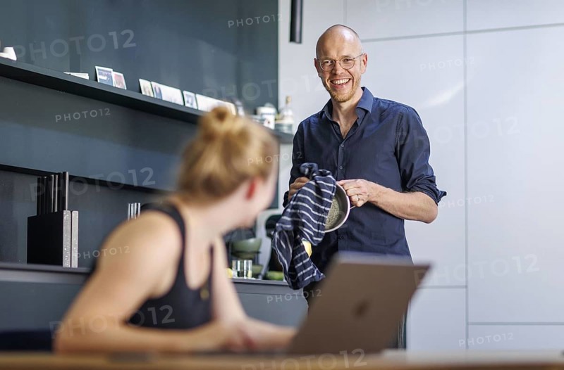 Symbolic photo on the subject of division of labour for couples in the household. A woman sits at a laptop in a kitchen while a man washes dishes in the background. Berlin, 13.08.2024