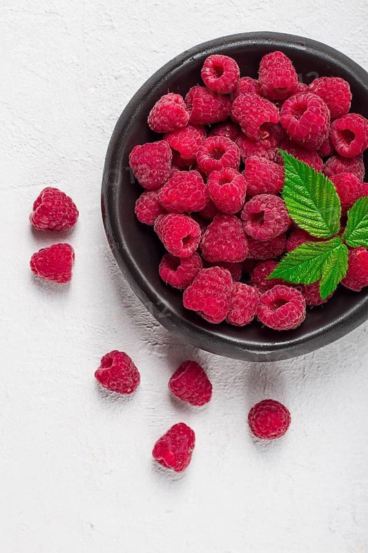 Fresh raspberries, on a black plate, with leaves, top view, on a white table
