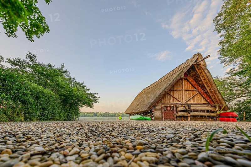 Thatched roof house next to a gravel path, surrounded by trees and grass under a cloudy sky, with canoes in the foreground, Stuttgart, Max-Eyth-See, Baden Würrtemberg, Germany, Europe