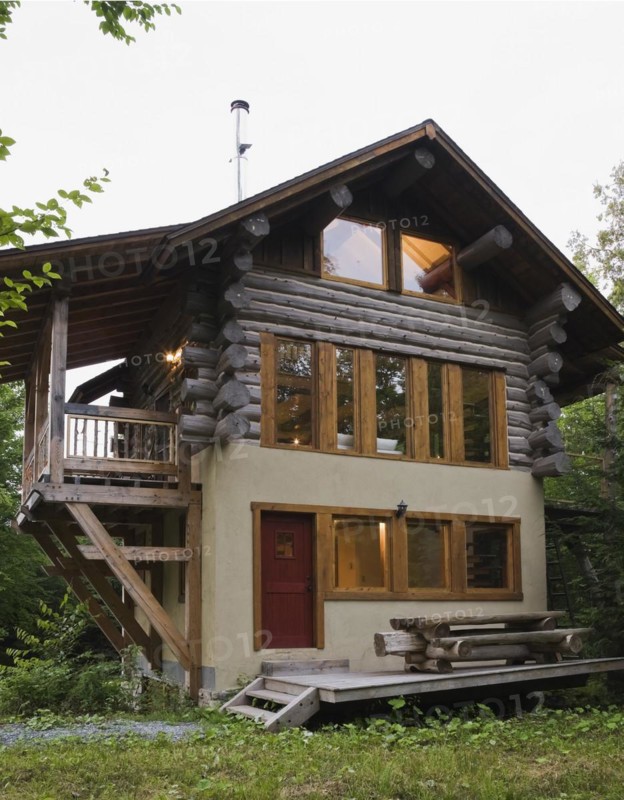 Three story Scandinavian hybrid style log cabin home facade cladded with beige stucco finish and timber wood balcony at dusk in summer, Quebec, Canada, North America