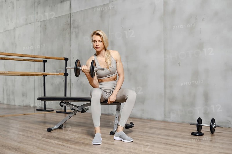 Muscular middle-aged woman sits on a bench in gym and lifting dumbbell while doing exercises for biceps, front view