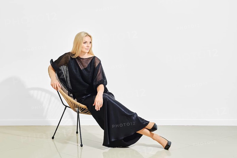 Calm and positive woman in black shiny dress lounging on a modern wicker chair in a bright room