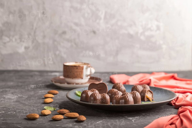 Chocolate caramel candies with almonds and a cup of coffee on a black concrete background and red textile. Side view, copy space