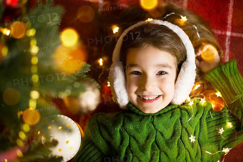 A happy girl in fur headphones lies near the Christmas tree in garlands and lights in a warm sweater on a cozy blanket. Christmas and New Year, holiday eve, magical mood