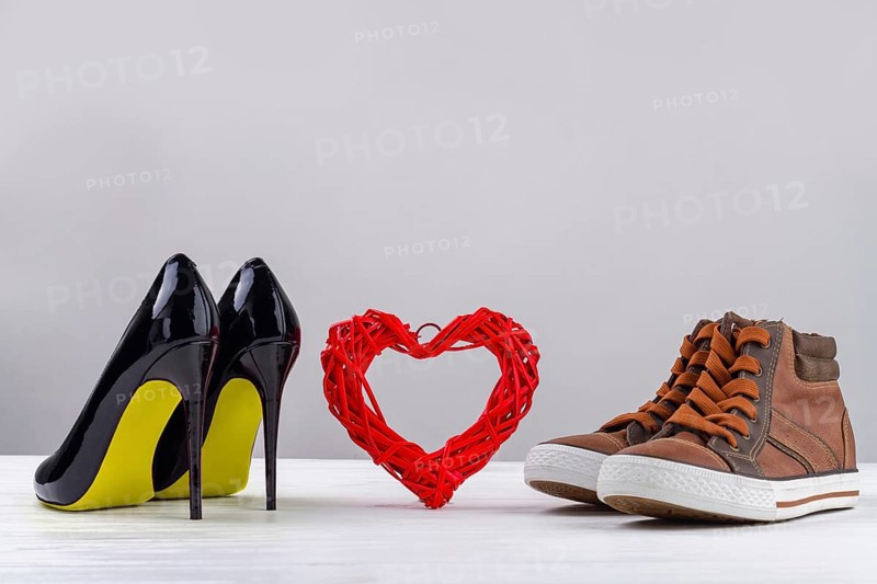 Red knitted heart with black womens high heel shoes and mens sneakers. Footwear on white background with copy space