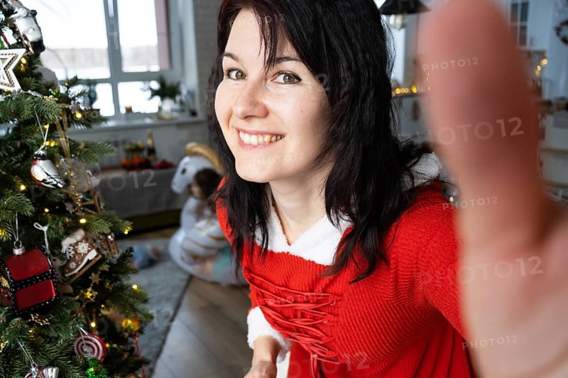 Selfie portrait of a woman in a red Santa dress in a home interior with a Christmas tree and New Year decor. Preparation for the holidays, party