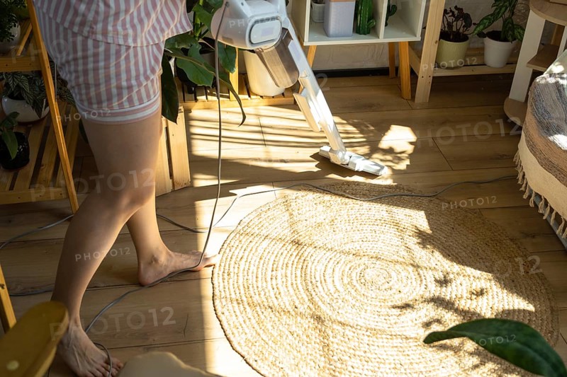 A woman vacuums a round carpet in a house among house plants with a hand vacuum cleaner. General cleaning of the house, cleaning service and housewife