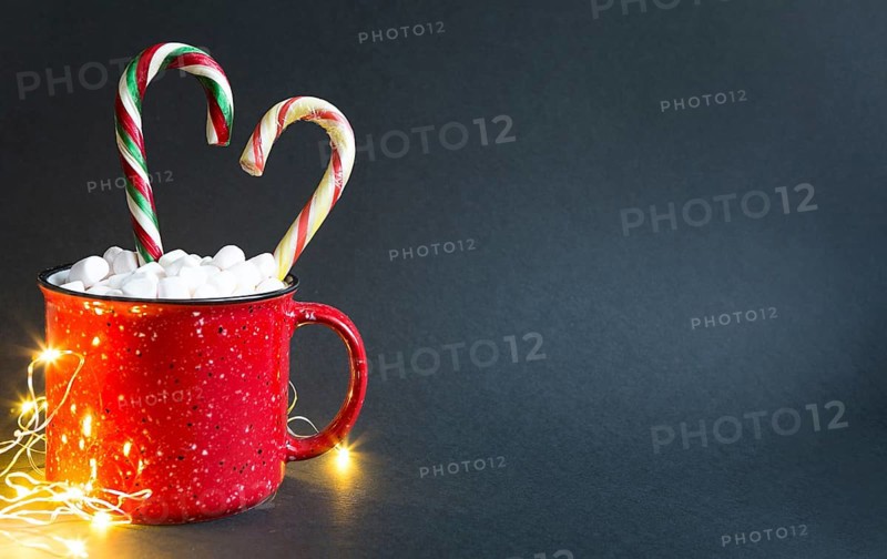 Red mug with marshmallows and caramel candy cane in the shape of a heart and lights of garlands on a black background. Christmas, New Year, festive mood. Copy space