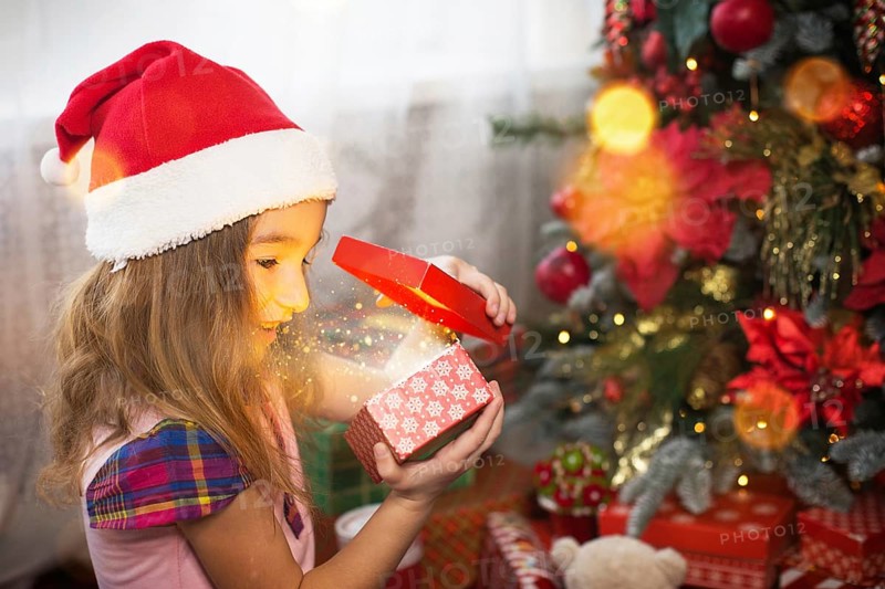 Little girl in Santa hat opens a red box with a gift and a Golden magic light near the Christmas tree. Holiday decor, poinsettias on fir trees, New year. Joy, surprise, children's emotions. Copyspace