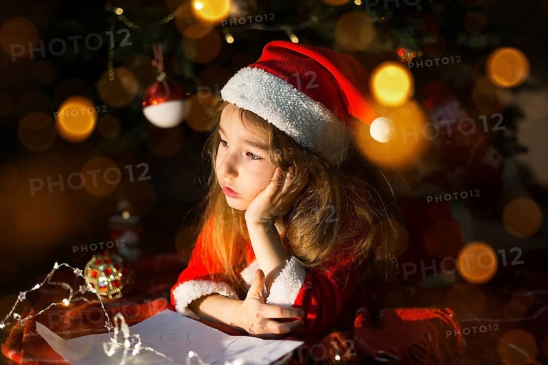 Little girl in a Santa hat and red dress under Christmas tree is dreaming, waiting for the holiday, lying on a plaid blanket. A letter on piece of paper, gifts. New year, Christmas. defocus lights