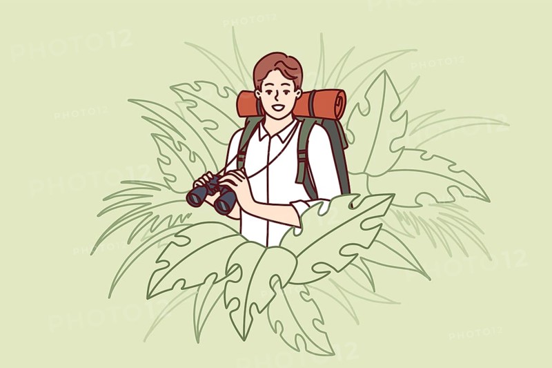 Man traveler stands in thickets of jungle and holds binoculars exploring area in search of further path. Guy traveler with backpack behind back loves adventure and is engaged in hiking