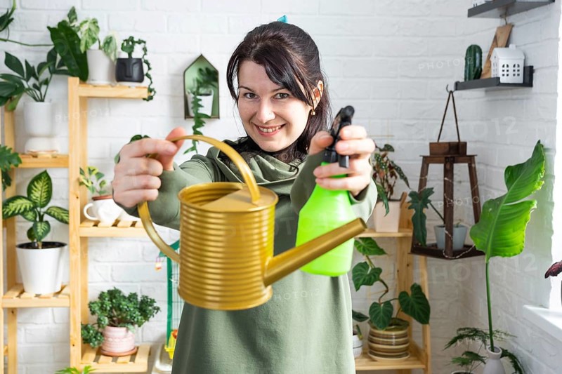 A portrait of a woman looks into the frame with an interior watering can and a spray gun for caring for home potted plants. Home plant growing, green house, water balance
