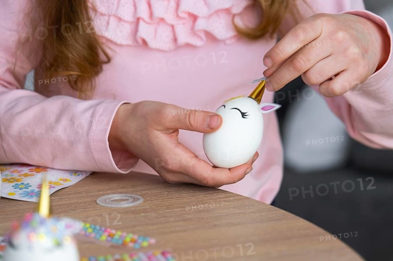 A cute girl with pink bunny ears makes an Easter craft, decorates an egg in the form of a unicorn with rhinestones, horn, flowers in the interior of a house with plants