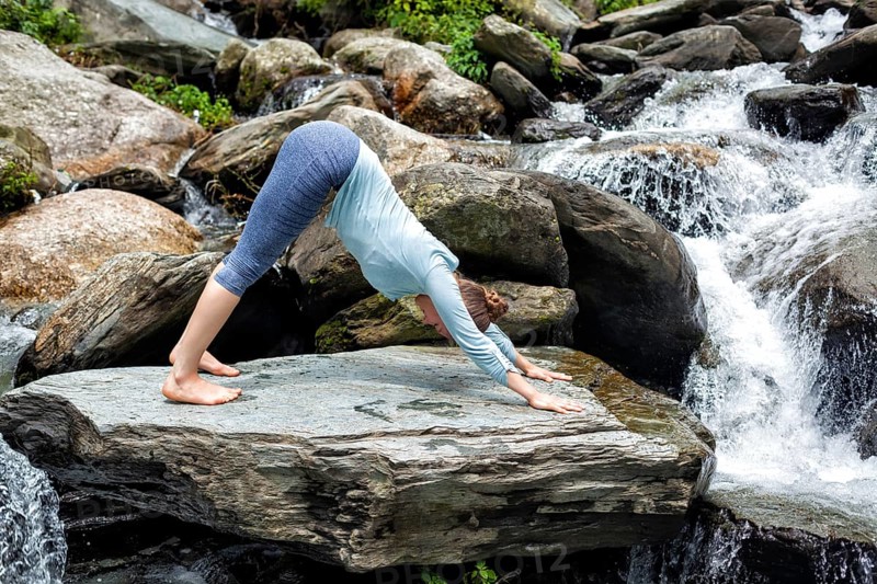 Young sporty fit woman doing yoga asana Adho mukha svanasana, downward facing dog, at tropical waterfall