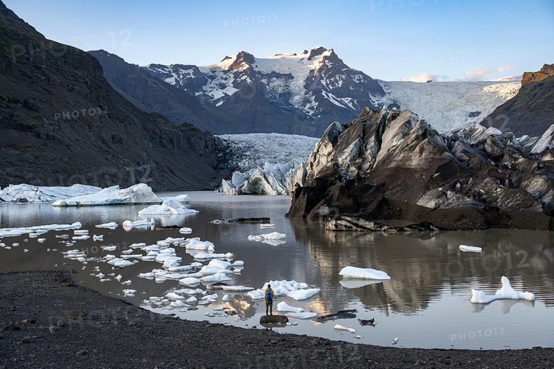 Hiker standing on a rock in the lake, reflection in the Svínafellslon glacier lagoon with ice floes, Svínasfellsjökull glacier tongue, glaciated mountain peak Hrútfjallstindar in the background, at sunset, Svínafell, Iceland, Europe