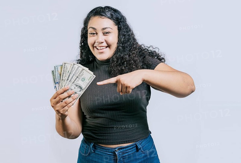 Attractive latin girl holding and pointing money. Smiling young woman holding and pointing money isolated. Latin people holding money