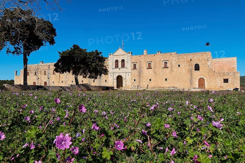 Wide angle, main side, gate entrance, trees, spring meadow, purple flowers, fortress-like walls, blue cloudless sky, Arkadi, Orthodox Monastery, National Monument, Rethimnon Province, Crete, Greece, Europe
