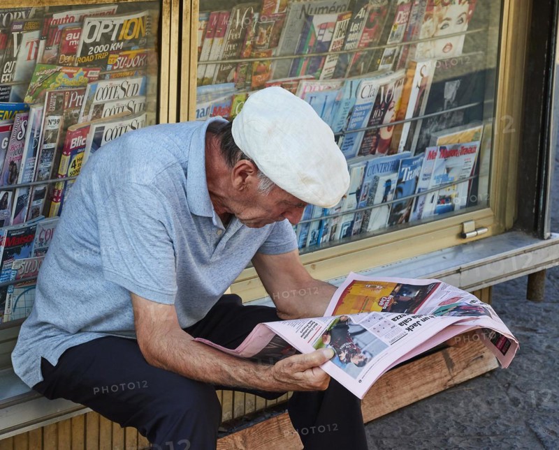 Older man sitting in front of kiosk reading a daily newspaper, white cap, old town, Catania, east coast, Sicily, Italy, Europe