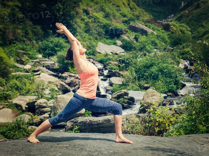 Yoga outdoors, sporty fit woman doing Ashtanga Vinyasa Yoga asana Virabhadrasana 1 Warrior pose posture at waterfall in HImalayas mountains. Vintage retro effect filtered hipster style image