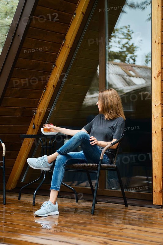 Woman sits in wicker chair near the table at wooden terrace of her forest cabin